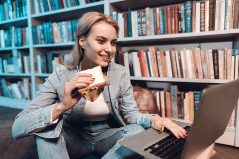 Young woman eating a sandwich as using her laptop in a library