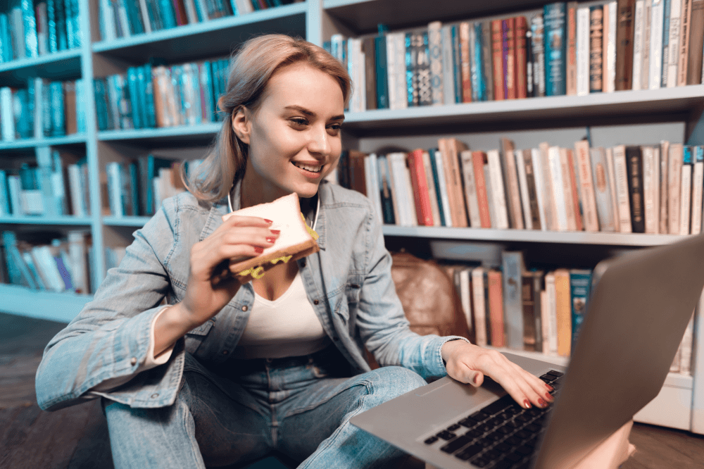 Young Woman Eating A Sandwich As Using Her Laptop In A Library