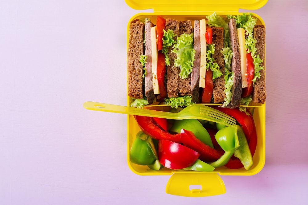 Healthy-school-lunch-box-with-beef-sandwich-fresh-vegetables-pink-table-top-view-flat-lay